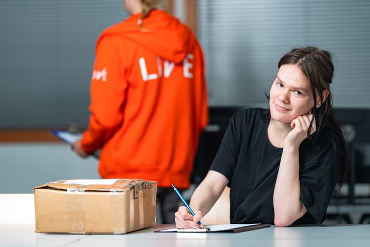 Opiskelija istuu pöydän ääressä kirjoittamassa muistivihkoon, vieressä pahvilaatikko. Toinen henkilö seisoo taustalla oranssissa hupparissa.