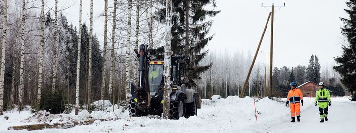 Metsätyökone työskentelee lumisessa metsässä, kaksi työntekijää neonvärisissä vaatteissa tarkkailee tilannetta.