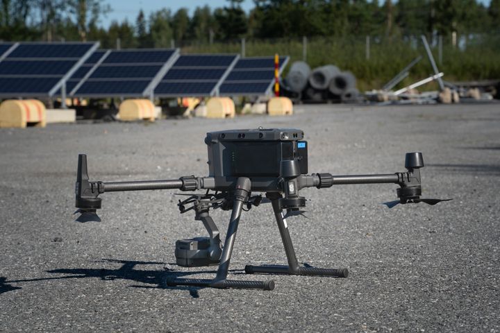 Neliroottorinen%20drone%20maassa%2C%20taustalla%20aurinkopaneeleita%20ja%20mets%E4%E4.