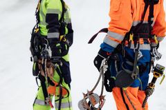 Kaksi pylvääseen kiipeävää asentajaa keltaisissa ja oransseissa työasuissa valjaissa ja työkaluvyö taskuineen ja kiipeilyvälineineen.