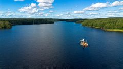 Lautta asentaa vesistökaapelia järven keskellä, taustalla näkyy metsäistä saarta ja rantaviivaa.