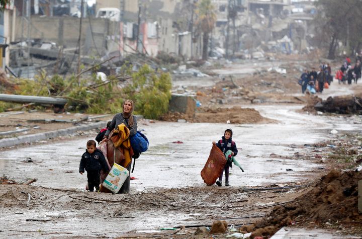 Äiti lasten kanssa kantaa tuskaisena painavia tavaroita ja kävelee tulvivalla ja mutaisella tiellä, taustalla pommitettuja rakennuksia