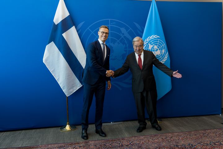 Suomen lippu ja YK:n lippu taustalla, presidentti ja pääsihteeri iloisesti kättelemässä