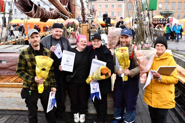 Voittajien poseeraus ulkona: kuusi henkilöä pitelee kukkakimppuja ja diplomeja tapahtumassa.