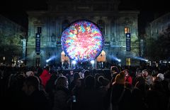 Kuva esittää suurta, värikästä, pallomaista taideteosta, joka hohtaa yöllisessä kaupunkimaisemassa ja on ihmisten ympäröimä.