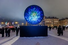 Alt-teksti: Valotaideteos ulkona talvisessa ympäristössä, valaistu sinisestä vihreään siirtyvillä väreillä. Yleisö seisoo teoksen ympärillä.