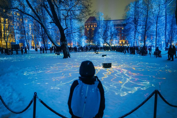 Henkilö katsoo lumista puistoa, jota valaisee värikäs valoteos. Puistossa on sininen valo.