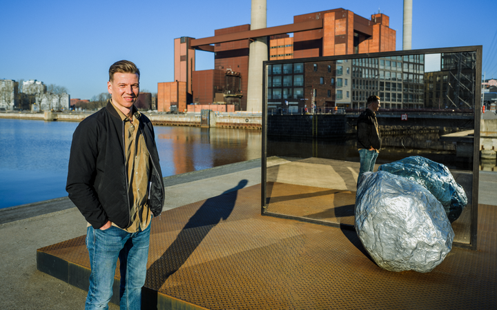 Sami Ylisaari seisoo nykyaikaisen teollisuusrakennuksen edessä veden äärellä. Lähellä on suuri peili ja hopeinen kivi.
