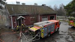 Palomiehet sammuttavat paloa punaisessa puutalossa. Paloautoja pihassa.