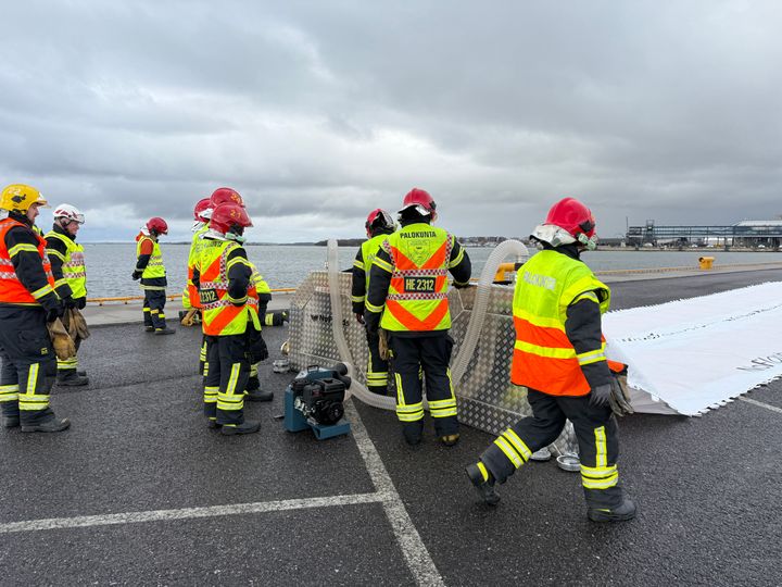 Palomiehet pystyttämässä vedellä täytettävää tulvapatoa Hernesaaren satama-alueella. Heillä on yllään kirkkaat pelastusasusteet ja kypärät. Taustalla näkyy pilvinen taivas ja meri.