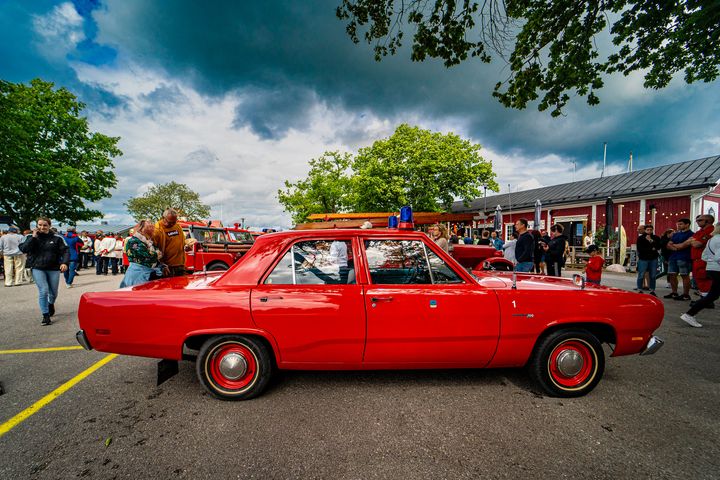 Punainen vuoden 1969 Plymouth Valiant palopäällikön auto pysäköitynä ulkona, ympärillä ihmisiä ja puita.