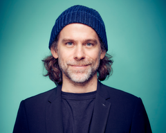 Bryce Dessner sinisessä pipossa ja puvuntakissa, vaaleansinisellä taustalla. Dessner hymyilee kuvassa.