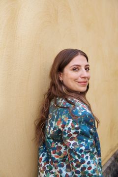 Dalia Stasevska seisoo vaaleankeltaisen seinän edessä värikkäässä takissa ja hymyilee olkansa yli.