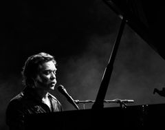 Rufus Wainwright esiintymässä mikrofonin äärellä flyygelin edessä. Kuva on mustavalkoinen.