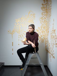 Nainen istuu tikkailla, kädessään maalia ja pensseli, taustalla seinätaidetta.