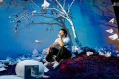 Näyttelijä istuu lavasteissa, joissa on paperia, koivupuu ja värikäs tausta.