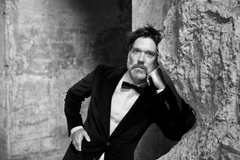 Rufus Wainwright pukeutuneena mustaan pukuun ja rusettiin. Hän nojaa karkeaan betoniseinään ja katsoo kameraan vakavana.