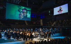 Orkesteri esiintyy konserttisalissa, jossa on kaksi suurta taustaprojektoria näytöillä.