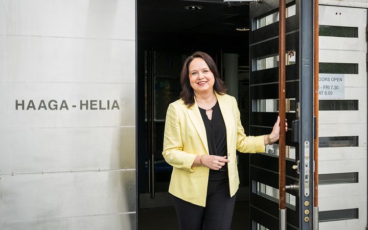 Susanna Niinistö-Sivuranta seisoo Haaga-Helia-kyltin vieressä ja pitää ovea auki. Hän hymyilee kameralle.