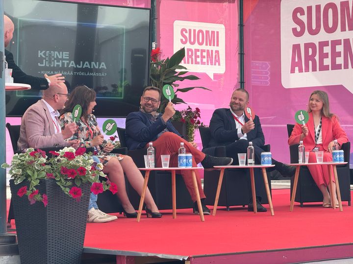 Viisi henkilöä istuu lavalla paneelikeskustelussa SuomiAreena-tapahtumassa. Heillä on käsissään kylttejä, ja he keskustelevat hymyillen.
