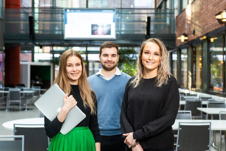 Kolme henkilöä seisoo sisätiloissa, taustalla näkyy suuri näyttö.