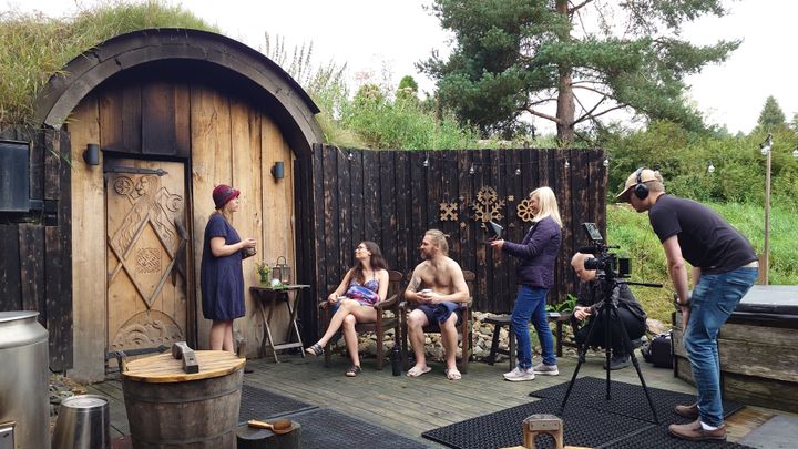 Ryhmää kuvataan saunan edustalla kesäisessä ympäristössä. Kaksi henkilöä istuu ja kolme seisoo.