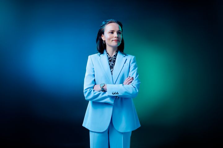 Henkilö vaaleansinisessä puvussa hymyilee ja poseeraa kädet ristissä sinivihreällä taustalla.