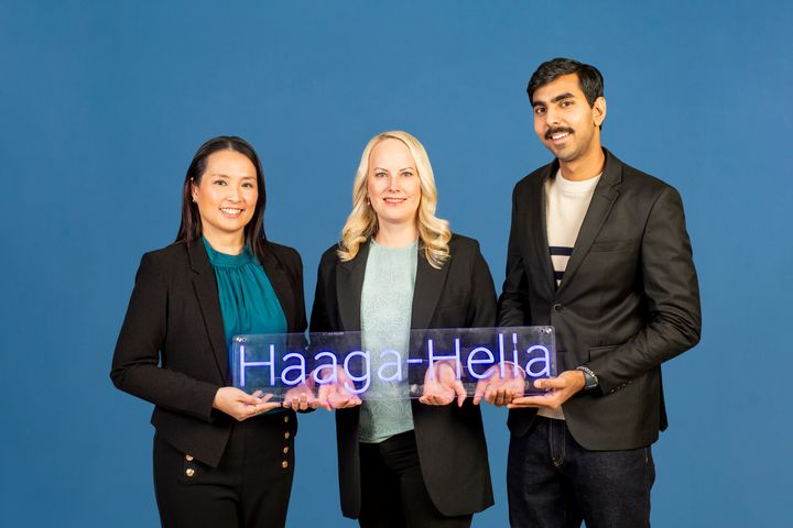 Kolme henkilöä seisoo sinistä taustaa vasten pitäen Haaga-Helia-kylttiä.