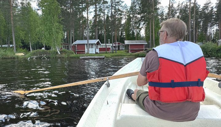 Mies soutaa venettä pelastusliivit päällään. Taustalla näkyy mökkirakennuksia.