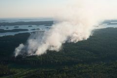 Kuva lintuperspektiivistä metsäpalosta. Paljon savua nousee metsästä. Helikopteri lentää savupatsaan edessä. Taustalla näkyy suomalainen järvimaisema.