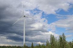 Kuva-alan vasemmassa reunassa valkoinen tuulivoimala, taustalla pilviä ja metsää.