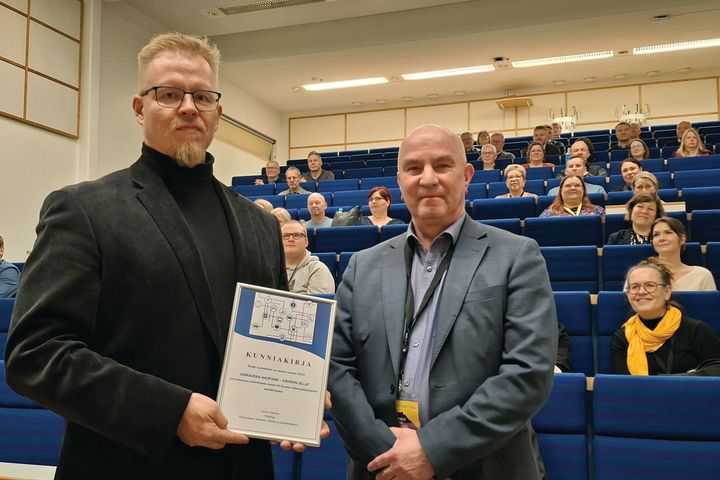 Kaksi palkittua miestä ottaa vastaan kunniakirjan auditoriossa.
