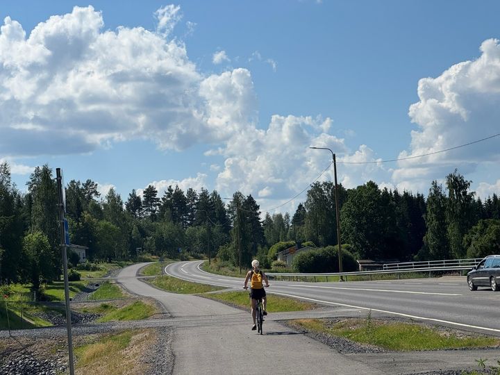 Nainen ajaa pyörällä poispäin kamerasta kesäisellä pyörätiellä.