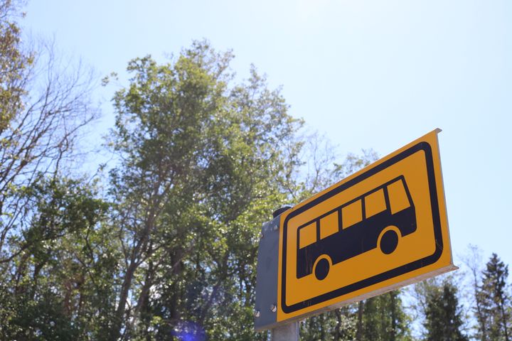 Linja-autopysäkin keltainen kyltti, taustalla puita ja sininen aurinkoinen taivas.