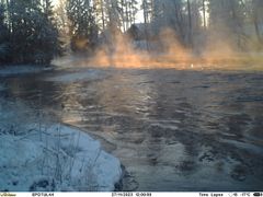 Öppet vatten där det ligger issörja. I framkanten syns den snöiga stranden. På andra sidan ån finns snöklädda träd och bland dem skymtar ett hustak.