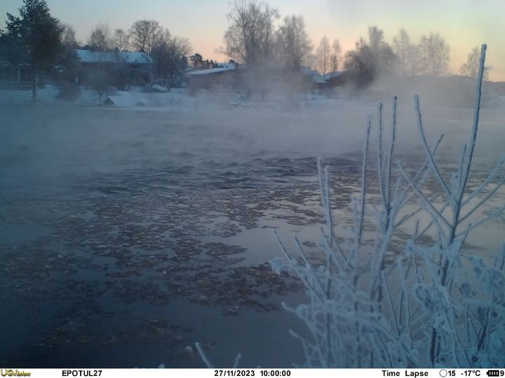 Öppet vatten i Esse å där det strömmar issörja. I framkanten finns frostiga småbjörkar och på andra sidan ån på den snöiga stranden finns några hus.