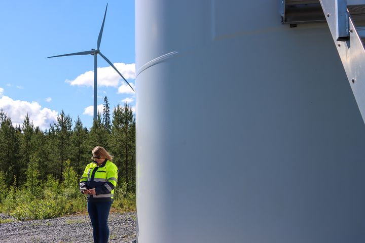 Henkilö seisoo tuulivoimalan lähellä. Taustalla näkyy metsä ja toinen tuulivoimala.