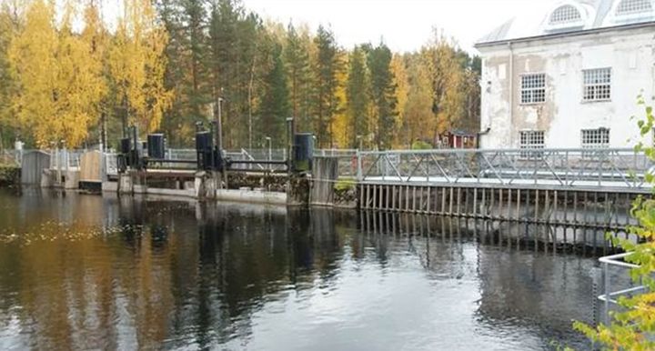 Voikosken patoallas, etualalla rauhallinen vesi, taustalla ruskaan siirtyviä puita ja vaalea voimalarakennus