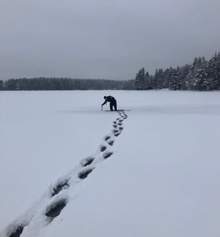 Jalan jäljet jäällä johtavat keskelle kuvaa, jossa mies mittaa jään paksuutta lumen läpi kairalla.