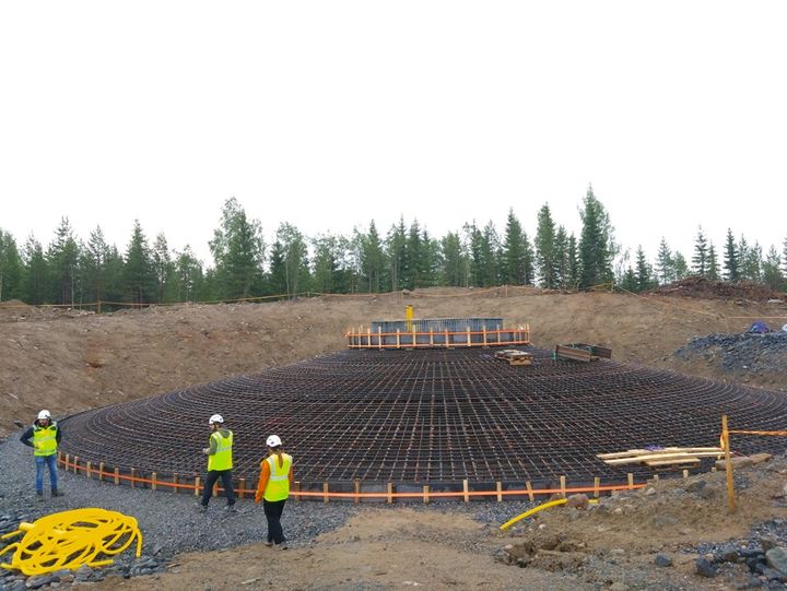 Rakennustyömaa, jossa on suuri teräsbetoniperustus. Kolme ihmistä, joilla on yllään huomioliivit ja kypärät, työskentelemässä. Taustalla näkyy pieni metsäalue ja etualalla on rakennusmateriaaleja ja -välineitä.