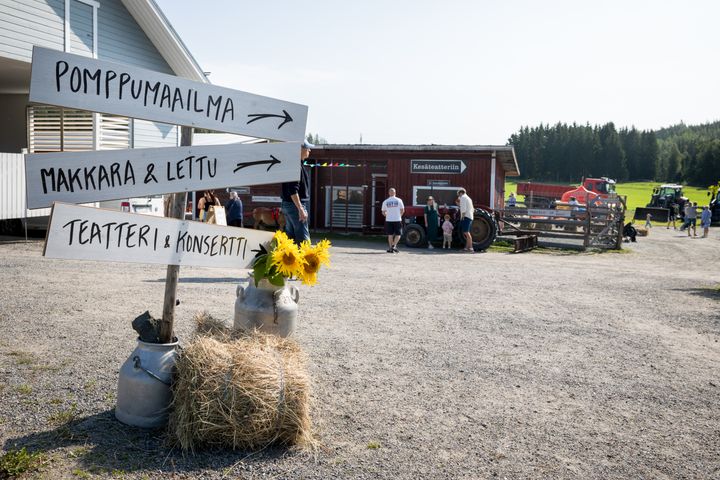 Kylttejä ohjeistamaan suuntaa, paali, kukka, taustalla punainen rakennus, ihmisiä ja traktoreita