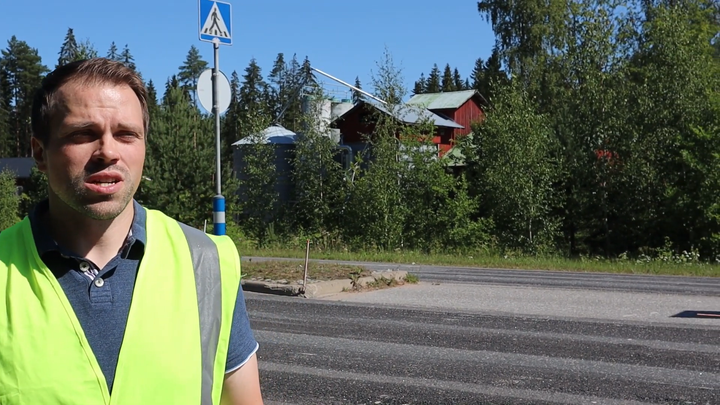 Kuvassa mies keltaisessa huomioliivissä seisoo suojatien vieressä kesäisessä maisemassa.