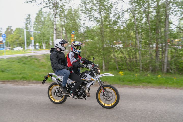 Kaksi henkilöä ajaa moottoripyörää maantiellä, ympärillä vehreä maisema.