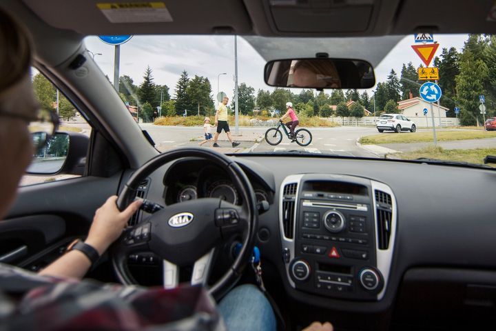 Kuljettajan näkymä autosta; suojatietä ylittää jalankulkija ja pyöräilijä.