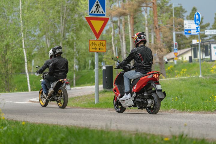 Kaksi motoristia ajamassa tiellä, ympärillä vihreää luontoa ja liikennemerkkejä.