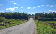 Maaseutumaisema, jossa pieni silta ylittää vesistön. Sillalla on liikennemerkkejä ja ympärillä metsää sekä avoin taivas.