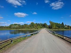 Maaseutumaisema, pitkä kapea silta ylittää rauhallisen järven. Molemmilla puolilla vehreää metsää ja sininen taivas, jossa on muutamia pilviä.