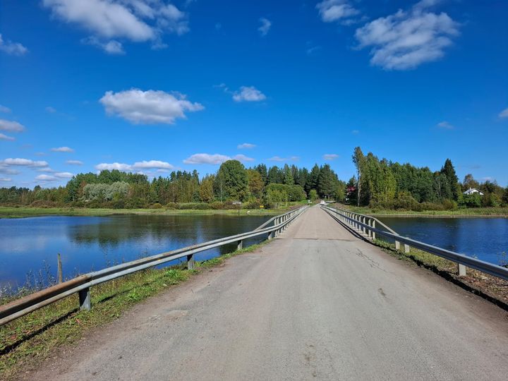 Maaseutumaisema, pitkä kapea silta ylittää rauhallisen järven. Molemmilla puolilla vehreää metsää ja sininen taivas, jossa on muutamia pilviä.