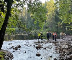 Ryhmä ihmisiä seisomassa joen varrella metsäisessä ympäristössä.