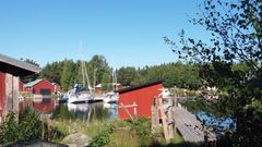 Kuvassa on Kaunissaaren idyllinen rantamaisema, jossa näkyy veneitä, punaisia mökkejä ja vehreää kasvillisuutta.